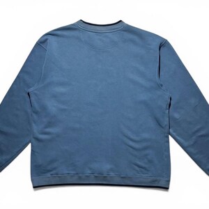 May include: A light blue sweatshirt with long sleeves, a crew neck, and a dark blue trim. The garment is made of a soft material and is laid flat on a white surface. The sweatshirt has a simple, classic design.