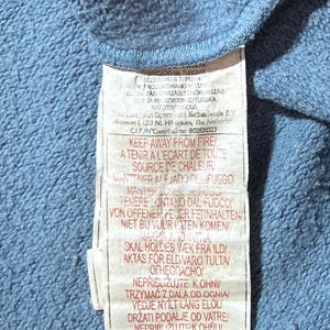 May include: A close-up of a blue fabric with a white tag attached. The tag has text in multiple languages, including English, French, and Spanish, with warnings about fire safety.