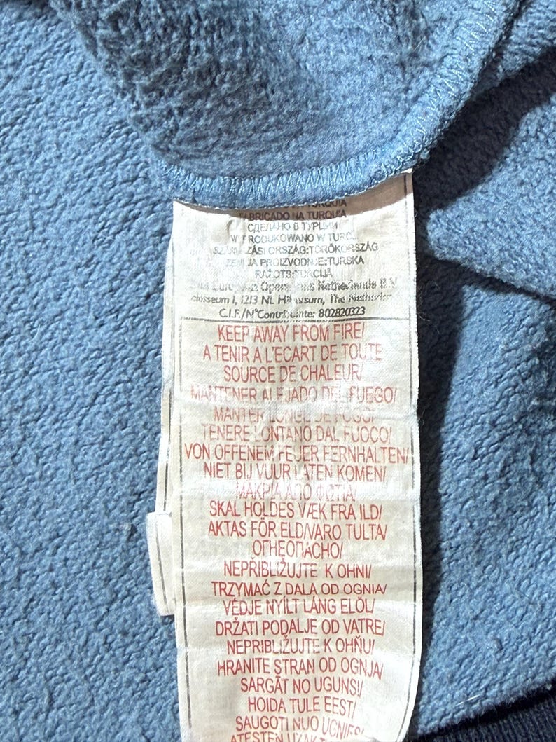 May include: A close-up of a blue fabric with a white tag attached. The tag has text in multiple languages, including English, French, and Spanish, with warnings about fire safety.
