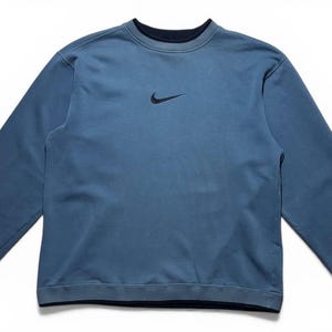 May include: A blue crew neck sweatshirt with long sleeves. The sweatshirt has a small black Nike logo on the chest. The collar, cuffs, and hem are a darker shade of blue. The garment is made of a soft material.