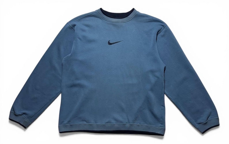 May include: A blue crew neck sweatshirt with long sleeves. The sweatshirt has a small black Nike logo on the chest. The collar, cuffs, and hem are a darker shade of blue. The garment is made of a soft material.