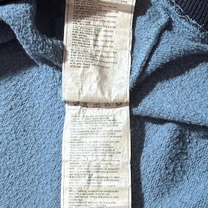 May include: Close-up of a blue and navy blue garment tag with multiple lines of text in various languages. The tag is attached to a blue fabric, likely part of a sweatshirt or similar clothing item.