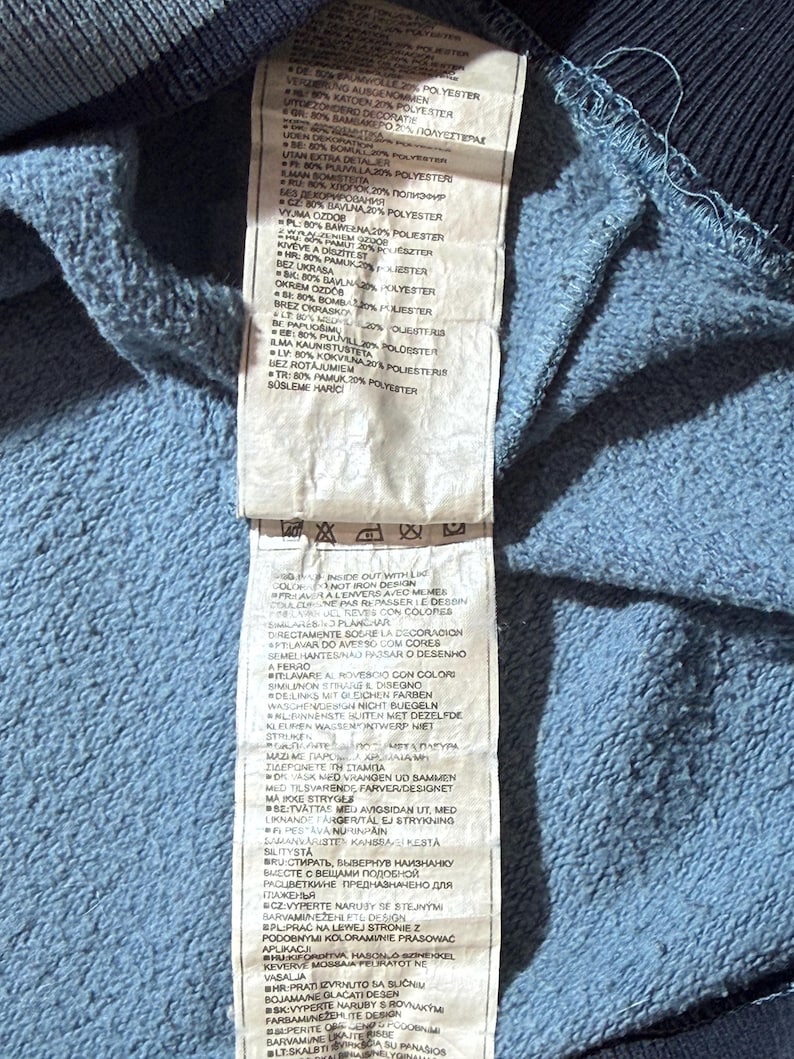 May include: Close-up of a blue and navy blue garment tag with multiple lines of text in various languages. The tag is attached to a blue fabric, likely part of a sweatshirt or similar clothing item.