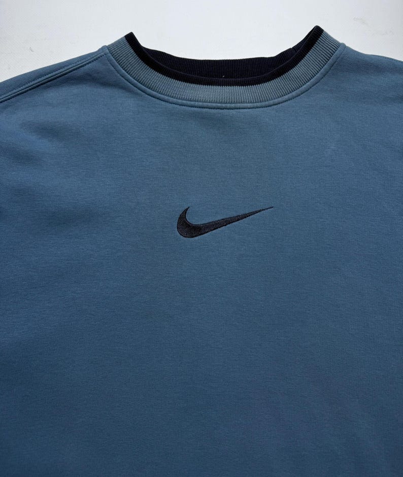 May include: A blue-grey sweatshirt with a black embroidered Nike swoosh logo on the chest. The crew neck has a black ribbed trim, and the cuffs and hem are solid. The sweatshirt is made of a soft, comfortable material.