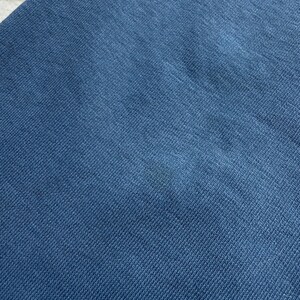 May include: Close-up of a section of blue fabric. The fabric has a textured appearance with a diagonal weave pattern. The color is a solid, medium shade of blue, with a slight variation in tone across the surface.