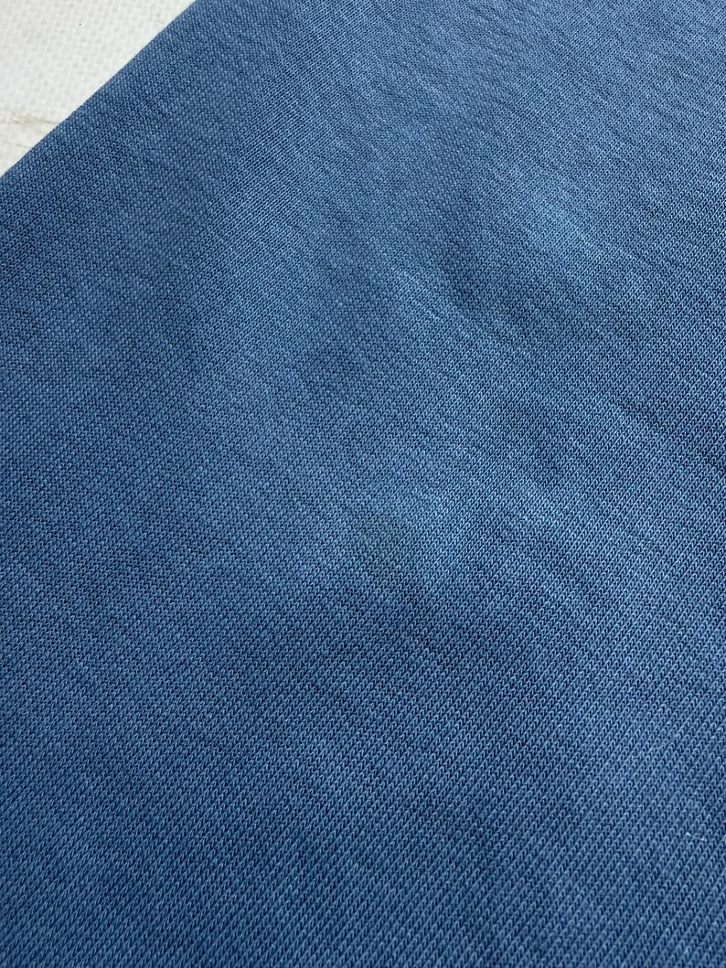 May include: Close-up of a section of blue fabric. The fabric has a textured appearance with a diagonal weave pattern. The color is a solid, medium shade of blue, with a slight variation in tone across the surface.