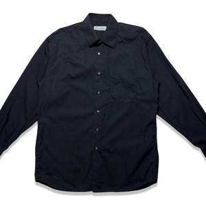 May include: A black long-sleeve button-down shirt with a classic collar and a chest pocket. The shirt has a subtle vertical stripe pattern and a small label near the collar. The shirt is spread out on a white surface.