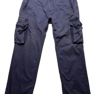 Può includere: Pantaloni cargo blu con più tasche sulle gambe. I pantaloni hanno una chiusura con bottone e cerniera, passanti per cintura e cinghie regolabili. Il tessuto sembra essere una resistente misto cotone. I pantaloni sono di colore blu.