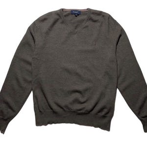 Può includere: Un maglione grigio scuro con scollo a V e maniche lunghe. Il maglione ha un orlo e polsini a coste. Un piccolo logo è visibile sul petto. Il maglione è realizzato in morbido tessuto a maglia.