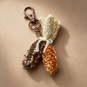 May include: A crochet keychain featuring two small, textured corn cobs attached to a bronze-toned metal lobster clasp. One cob is variegated with brown, green, and cream yarn, while the other is orange and yellow. A cream yarn bow ties the cobs together.