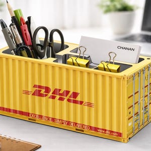 Peut inclure: Organisateur de bureau jaune en forme de conteneur maritime, avec le logo DHL en rouge. Il contient des stylos, des ciseaux et des cartes de visite. Le texte "EXCELLENCE. SIMPLY DELIVERED." est imprimé.