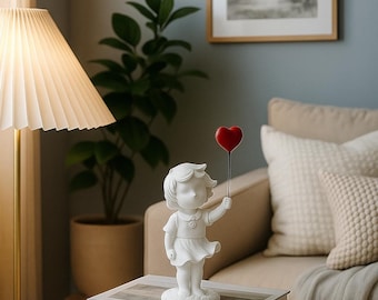 May include: A white figurine of a girl holding a red heart-shaped balloon. The statue is on a stack of books on a wooden side table. A lamp and a framed picture are in the background.