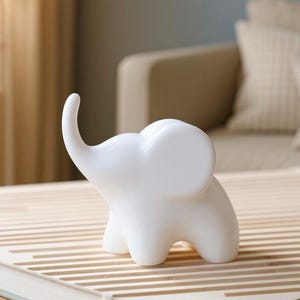 May include: A small, white ceramic elephant figurine with a raised trunk and large ears. The elephant is positioned on a light-colored, striped surface, likely a book or decorative tray. The background is softly blurred, suggesting a living room setting.