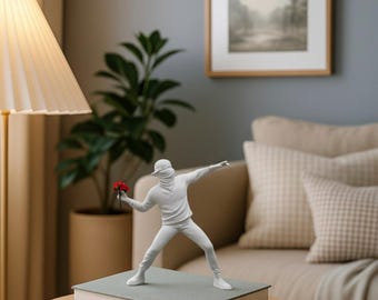 May include: A white sculpture of a figure throwing a bouquet of red flowers, standing on a stack of books. The figure is wearing a cap and a face covering. The sculpture is on a round wooden table, with a lamp and a sofa in the background.