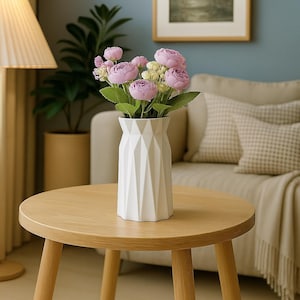 Puede incluir: Un jarrón blanco geométrico con flores rosas y amarillas sobre una mesa redonda de madera. El jarrón tiene un diseño moderno con facetas anguladas. La mesa está sostenida por tres patas de madera. El fondo incluye un sofá y un cuadro enmarcado.