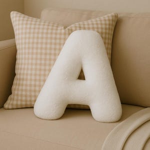 Peut inclure: Un coussin en forme de lettre "A" blanc et moelleux repose sur un canapé beige, avec un coussin à motif vichy derrière lui. Le coussin en forme de lettre a un aspect doux et texturé. L'arrière-plan comprend un canapé aux tons neutres et une couverture pliée.