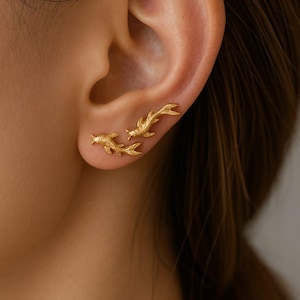 Peut inclure: Boucles d'oreilles dorées en forme de poissons. Les boucles d'oreilles sont portées sur l'oreille, avec un poisson près du lobe et deux poissons qui remontent l'oreille. Elles ont une surface texturée et une finition brillante.
