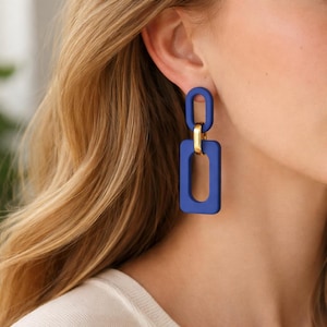 Peut inclure: Gros plan sur une boucle d'oreille géométrique bleue avec une forme rectangulaire et un connecteur doré. La boucle d'oreille est suspendue à l'oreille. C'est un accessoire audacieux et moderne.