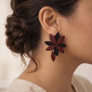 Peut inclure: Une boucle d'oreille imposante au design floral superposé, composée de pétales noir mat et bordeaux foncé. Le bijou est formé de plusieurs éléments en forme de pétales qui se chevauchent en étoile, avec un pétale suspendu vers le bas.