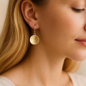 May include: Gold-coloured hammered disc earrings. The earrings are round with a textured surface and hang from a simple hook. The earrings are shown on a person with blonde hair.