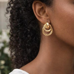 Könnte beinhalten: Goldfarbene Ohrringe mit einem Doppelreifen-Design. Der obere Reifen ist glatt, während der untere Reifen ein strukturiertes, geflochtenes Muster aufweist. Die Ohrringe werden an einem Ohr gezeigt, um ihre Größe und ihren Stil hervorzuheben.