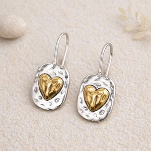 Peut inclure: Une paire de boucles d'oreilles argentées avec une texture martelée et un cœur doré au centre. Les boucles d'oreilles ont un design simple en forme de crochet. Les bijoux sont présentés sur un fond neutre.