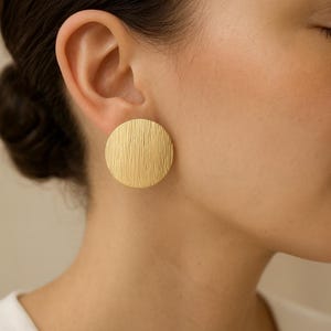 Op de afbeelding: Een close-up van een goudkleurige, ronde oorbel met een getextureerd oppervlak. De oorbel heeft de vorm van een grote schijf en wordt op het oor gedragen. De oorbel is een eenvoudig, maar elegant sieraad.