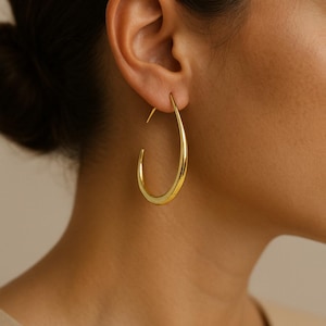 May include: A close-up of a gold-colored hoop earring. The earring has a teardrop shape and is worn on the ear. The earring is smooth and shiny, reflecting the light. The background is a neutral color.