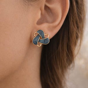 May include: A close-up of a gold-colored stud earring with a dark teal enamel design. The earring has a unique, intertwined shape, and is worn on a person's ear. The earring's design is a modern take on a classic style.
