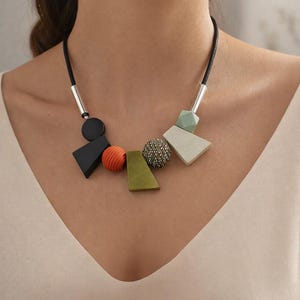 Peut inclure: Un collier moderne avec un cordon noir et des formes géométriques. Le collier comprend un triangle noir, une sphère orange côtelée, un trapèze vert, une sphère texturée et un trapèze gris. Des tubes métalliques argentés relient le cordon au pendentif.