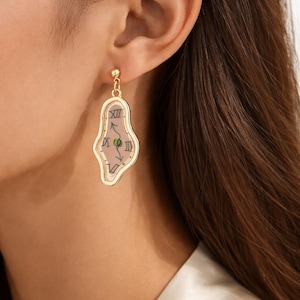 May include: Gold-toned drop earrings featuring a surrealist melting clock design. The earrings have a light pink background with black clock hands and Roman numerals. The earrings are a unique accessory.