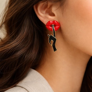 Peut inclure: Boucles d'oreilles pendantes avec une bouche rouge et une main noire pointant vers le haut. Les boucles d'oreilles ont un contour doré et sont portées sur l'oreille. Accessoire audacieux et tendance.