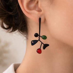 Puede incluir: Pendientes abstractos negros con un diseño lineal. Los pendientes presentan un tallo negro con acentos coloridos, que incluyen un círculo rojo, un círculo verde, una forma de hoja negra, un círculo azul y un círculo negro.
