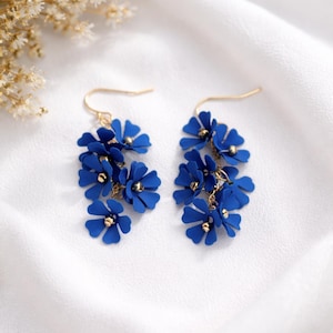 May include: A pair of dangling earrings featuring clusters of vibrant blue flowers with gold-colored accents. The earrings have gold-colored hooks and are set against a white background, creating a delicate and feminine aesthetic.