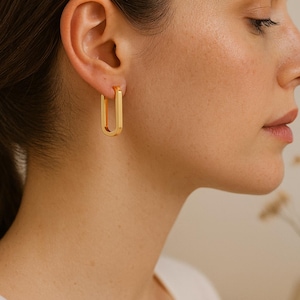 Peut inclure: Boucles d'oreilles créoles rectangulaires de couleur or. Les boucles d'oreilles présentent un design simple et moderne, avec une finition polie. L'image montre un gros plan des boucles d'oreilles sur l'oreille d'une personne.