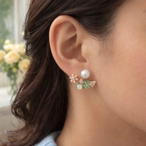 May include: A floral ear jacket earring with a large white pearl stud and a decorative floral design. The floral design features pink, green, and pastel-colored flowers and crystals. The earring is gold-toned.