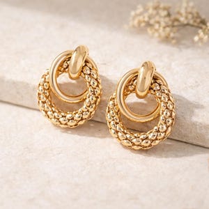 Peut inclure: Une paire de boucles d'oreilles dorées. Chaque boucle d'oreille présente un motif ovale texturé avec un anneau plus petit et lisse à l'intérieur. Une barre incurvée relie les deux anneaux. Les boucles d'oreilles sont présentées sur une surface claire.