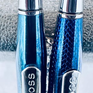 May include: Two blue Cross pens with silver accents are displayed. One pen has a smooth finish, while the other features a textured pattern. The word "CROSS" is engraved on each pen's silver clip. The pens are set against a textured gray background.