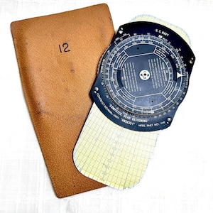 May include: A vintage U.S. Navy "Computer Dead Reckoning" navigation tool with a circular slide rule and a yellow and white grid. It is accompanied by a brown leather case with the number "12" on it.