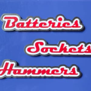 May include: Three rectangular signs with the words "Batteries", "Sockets", and "Hammers" in red, outlined in white and black. The signs are arranged on a blue background.