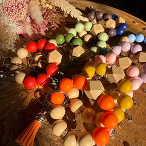May include: A collection of colorful wooden bead keychains in various colors, including red, orange, yellow, green, and blue. Some keychains feature geometric wooden beads. An orange tassel keychain is also visible. The keychains are arranged on a wooden surface.