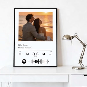 May include: Black framed print showing a couple on a beach at sunset. The print includes a music player interface with the song "Billie Jean" by Michael Jackson. The frame is black, and the print is displayed on a white surface.