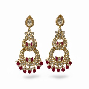 May include: A pair of gold-coloured chandelier earrings. Each earring has a teardrop-shaped top, followed by clear, round stones. Red and white beads dangle from the bottom of each earring.