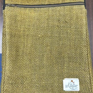 May include: A rectangular olive green laptop sleeve featuring a textured herringbone weave pattern. The sleeve includes a black zippered closure across the top and a small white rectangular fabric label in the bottom corner with the text Kiteflier.