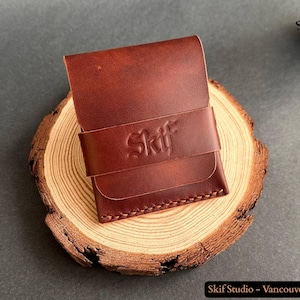 Peut inclure: Un portefeuille en cuir marron avec une fermeture à rabat et une sangle estampillée du mot "Skif". Le portefeuille est posé sur une tranche de bois, mettant en valeur sa couleur et sa texture riches. Les coutures sont visibles.