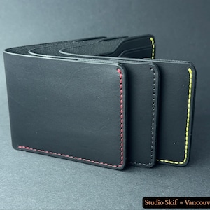 May include: Three black leather wallets with contrasting stitching. One wallet has red stitching, another has black stitching, and the third has yellow stitching. The wallets are stacked, showcasing their design and craftsmanship.