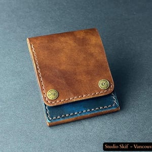 May include: A brown and blue leather wallet. The top flap is a warm brown, fastened with two brass-toned rivets. The lower section is a deep blue, with visible stitching. The wallet is closed and appears to be empty.