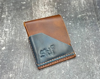 May include: A brown and blue leather wallet with the word "Skif" embossed on the front. The wallet has a bi-fold design with a blue pocket and brown leather exterior. The edges are stitched with brown thread and a blue accent.