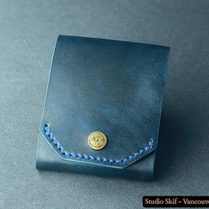 Peut inclure: Un portefeuille en cuir bleu foncé avec une fermeture à bouton en laiton et des coutures bleues. Le portefeuille rectangulaire semble plié. Le texte "Studio Skif - Vancouver, Canada" est en bas.
