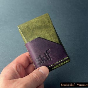 May include: A handmade leather wallet in olive green and purple. The wallet has a purple pocket with the word "Skif" embossed on it. The stitching is green. The wallet is held in a hand.